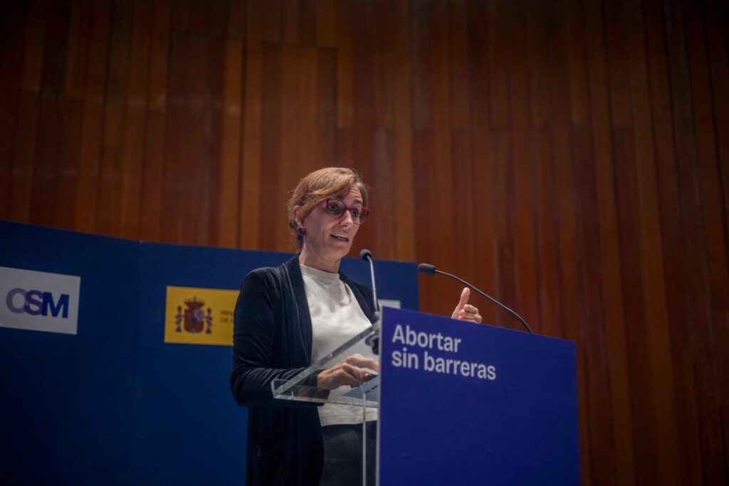 Una mujer habla en un atril con un cartel que dice "Abortar sin barreras", en un ambiente de conferencia.