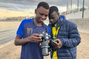 Dos jóvenes en una playa, mirando una cámara de fotos y un teléfono móvil, con edificios al fondo en Alcalá de Henares.