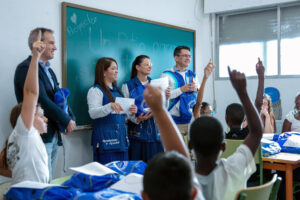 Entregan kits de material escolar en un aula de Torrejón de Ardoz, con adultos y niños con las manos levantadas.