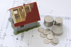 La imagen muestra una maqueta de una casa con el techo incompleto sobre planos arquitectónicos, junto a varias monedas apilad