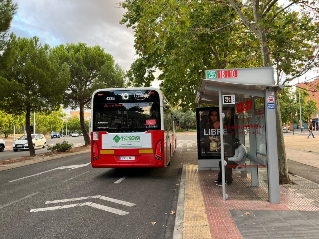 Autobús de la línea 10 en parada con un pasajero sentado bajo una marquesina en Alcalá de Henares, árboles y coches al fondo.