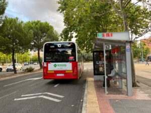 Autobús de la línea 10 en parada con un pasajero sentado bajo una marquesina en Alcalá de Henares, árboles y coches al fondo.