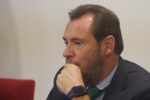 Hombre con barba y cabello castaño, pensativo, sentado en una mesa con fondo blanco y una silla roja, en Madrid.