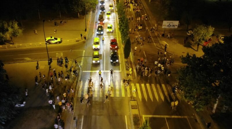 Grupo de personas cruzando un paso peatonal iluminado por luces de coches en Getafe, con vehículos detenidos y movimiento en