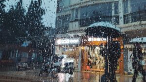 Imagen tomada a través de un cristal mojado, mostrando gotas de agua y siluetas de personas que sostienen paraguas mientras c